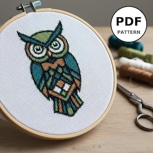 Peut inclure: Un motif de broderie au point de croix représentant un hibou dans un cercle à broder en bois. Le hibou présente des motifs géométriques bleu sarcelle, verts et bruns. Le texte "PDF PATTERN" est dans un cercle blanc. Fils à broder et ciseaux en arrière-plan.