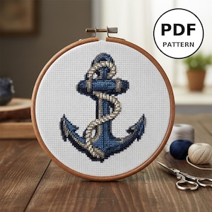 May include: A cross-stitch embroidery of a blue anchor with a rope detail, set in a wooden hoop. The design is on white fabric. The words "PDF PATTERN" are in a black circle in the upper right corner. The background is a wooden table.