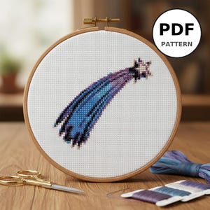 Peut inclure: Un motif de point de croix d'une étoile filante bleue et violette est présenté dans un cercle à broder en bois. Le texte "PDF PATTERN" est dans un cercle blanc. Des ciseaux dorés et des bobines de fil sont sur une surface en bois.