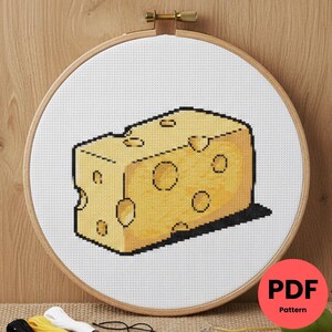 Peut inclure: Un motif de point de croix d'un bloc de fromage jaune avec des trous, présenté dans un cercle à broder en bois. Le fromage a une ombre, et le fond est blanc. Un cercle rouge avec le texte "PDF Pattern" est en bas à droite.