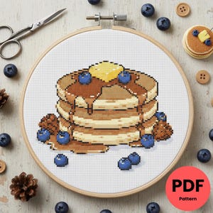 Peut inclure: Motif de point de croix d'une pile de crêpes avec du sirop, des myrtilles et une noix de beurre. Le motif est dans un cercle à broder en bois. Des myrtilles, des pommes de pin, des boutons et des ciseaux sont éparpillés autour du cercle.