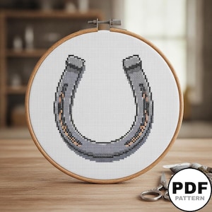 Peut inclure: Un motif de point de croix représentant un fer à cheval gris avec des contours noirs et des accents brun clair, présenté dans un tambour à broder en bois circulaire sur un tissu blanc. Le motif est centré dans le tambour avec des détails pixélisés.