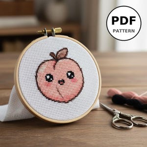 Peut inclure: Un motif de point de croix d'une pêche souriante dans un cercle à broder en bois. La pêche est rose avec une tige marron et des yeux noirs. Le texte "PDF PATTERN" est dans un cercle blanc. Ciseaux et fil sont également visibles.