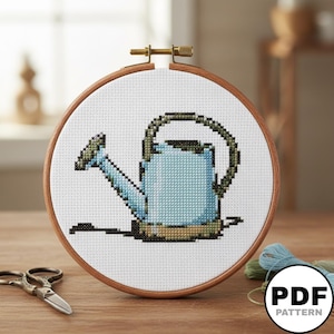May include: A cross-stitch project in a round wooden embroidery hoop featuring a light blue watering can with dark green accents on white fabric. The design includes a small black shadow at the base. A PDF pattern label is visible in the corner.