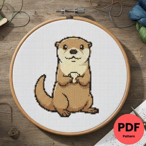 Peut inclure: Un motif de point de croix d'une loutre de dessin animé dans un cercle à broder en bois. La loutre est marron et beige, avec une expression souriante. Le motif est sur tissu blanc. Un cercle rouge avec "PDF Pattern" est visible.