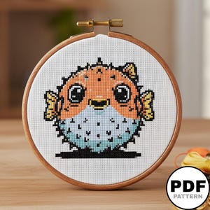 Peut inclure: Un projet de point de croix dans un tambour à broder rond en bois représentant un poisson-globe pixélisé. Le poisson a un corps orange rond, de grands yeux noirs et un ventre bleu clair avec de petites épines noires, brodé sur du tissu blanc.