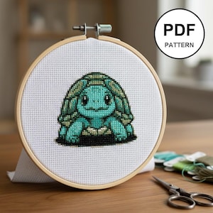 May include: A cross-stitch pattern of a cartoon turtle in a wooden embroidery hoop. The turtle is primarily teal and green, with a black base. The text "PDF PATTERN" is in a white circle in the upper right corner.