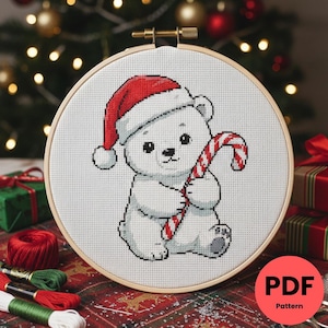 Puede incluir: Patrón de punto de cruz de un oso polar con un gorro de Papá Noel y un bastón de caramelo. El diseño está en un bastidor de bordado de madera. El oso es blanco con ojos y nariz negros. El gorro y el bastón tienen rayas rojas y blancas. El fondo es navideño.