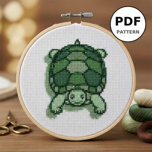 Peut inclure: Un motif de point de croix d'une tortue dans des tons de vert, présenté dans un cercle à broder en bois. Le motif est sur tissu blanc, avec le texte "PDF PATTERN" dans un cercle noir. Ciseaux et bobines de fil visibles.
