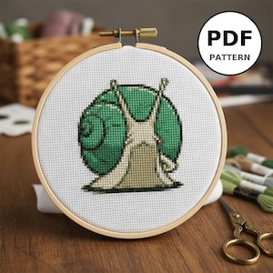 Peut inclure: Un cercle de broderie au point de croix avec un motif d'escargot vert et crème. L'escargot est sur un tissu blanc dans un cercle en bois. Le texte "PDF PATTERN" est dans un cercle blanc. Fils à broder et ciseaux en arrière-plan.