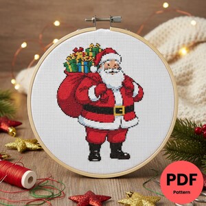 Peut inclure: Broderie au point de croix du Père Noël portant un sac de cadeaux, encadrée dans un cercle en bois. Le Père Noël est vêtu de son costume rouge traditionnel avec une ceinture et des bottes noires. Le fond est blanc, et le motif est étiqueté "PDF Pattern".