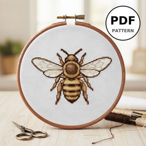 Peut inclure: Un motif de point de croix d'une abeille, présenté dans un cercle à broder en bois. L'abeille est réalisée avec du fil marron et jaune, et le cercle est placé sur un fond doux. Le texte "PDF PATTERN" est dans un cercle.