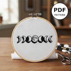 Peut inclure: Un motif de point de croix des phases de la lune, présenté dans un cercle à broder en bois. Le motif est brodé en fil noir sur du tissu blanc. Une étiquette PDF PATTERN est dans le coin supérieur droit. Ciseaux et fil à broder sont visibles.