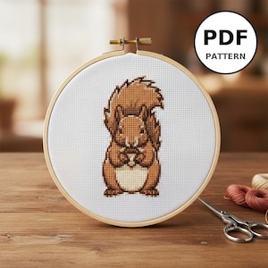 Peut inclure: Un motif de point de croix d'un écureuil tenant une noix, présenté dans un cercle à broder en bois. L'écureuil est brun et beige, avec une queue touffue. Le cercle est sur une surface en bois, avec des ciseaux et du fil à proximité. Le texte "PDF PATTERN" est dans un cercle blanc.