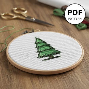Peut inclure: Un cercle de broderie au point de croix avec un motif d'arbre vert. L'arbre est brodé sur du tissu blanc et placé dans un cercle en bois. Des ciseaux dorés et du fil vert sont en arrière-plan. L'image comprend une icône de modèle PDF.