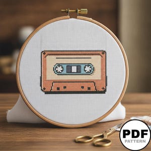 Peut inclure: Un projet de broderie au point de croix représentant une cassette audio pixelisée orange et bleue sur tissu blanc, fixé dans un tambour à broder circulaire en bois avec une vis de tension en métal, présenté comme un modèle PDF numérique.