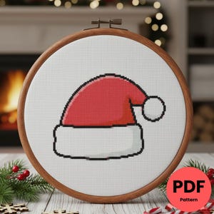 Peut inclure: Un motif de point de croix d'un bonnet de Père Noël dans un cercle à broder en bois. Le bonnet est rouge avec un bord blanc et un pompon blanc. Le fond est blanc et le cercle est marron. Un cercle rouge avec "PDF Pattern" est en bas à droite.