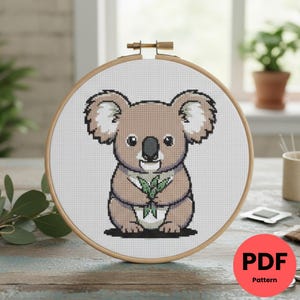 Peut inclure: Un motif de point de croix d'un koala tenant des feuilles d'eucalyptus, présenté dans un cercle à broder en bois. Le koala est brun clair avec des détails blancs et un nez noir. Le motif est sur tissu blanc. Un cercle rouge avec "PDF Pattern" est en bas à droite.