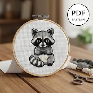 May include: A cross-stitch embroidery hoop with a grey and white raccoon design. The raccoon is on white fabric within a wooden hoop. The text "PDF PATTERN" is in a black circle in the upper right corner.