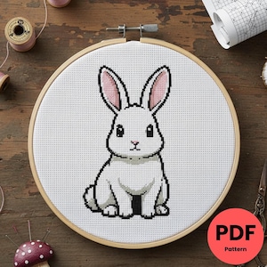 Peut inclure: Un motif de point de croix d'un lapin blanc avec l'intérieur des oreilles roses, encadré dans un cercle à broder en bois. Le lapin est centré sur un fond de tissu blanc. L'image comprend une étiquette de motif PDF.