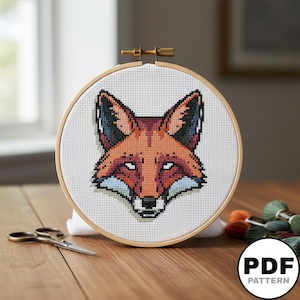 Peut inclure: Un tambour à broder circulaire en bois contenant un tissu Aida blanc avec un motif au point de croix représentant une tête de renard. Le renard présente des fils orange, rouges, noirs et blancs. Des ciseaux en métal sont posés à gauche.