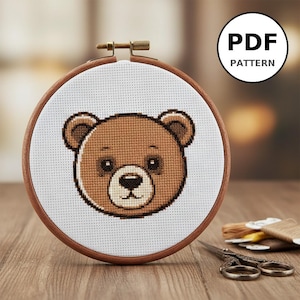 Peut inclure: Un cercle de broderie au point de croix avec un motif d'ours en peluche marron sur tissu blanc. L'ours a des yeux marron foncé et un museau brun clair. Le cercle en bois est fixé avec une vis en laiton. Le texte "PDF PATTERN" est dans un cercle blanc.