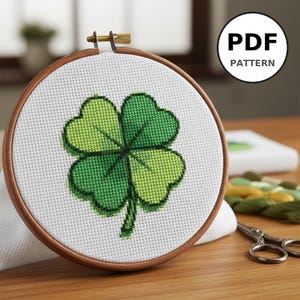 Peut inclure: Un cercle de broderie au point de croix avec un motif de trèfle à quatre feuilles dans des tons de vert. Le motif est posé sur un fond de tissu blanc, encadré par un cercle en bois. Le texte "PDF PATTERN" est dans un cercle blanc.