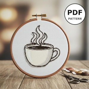 Peut inclure: Un motif de point de croix d'une tasse de café avec de la vapeur, présenté dans un cercle à broder en bois. Le motif est en noir et blanc, avec le texte "PDF PATTERN" dans un cercle. Le cercle est sur une surface en bois.