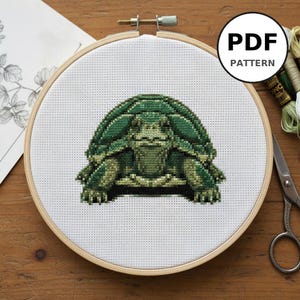 Peut inclure: Un motif de point de croix d'une tortue dans des tons de vert, présenté dans un cercle à broder en bois. Le motif est sur du tissu blanc, avec le texte "PDF PATTERN" dans un cercle blanc. Ciseaux et fil à broder sont visibles.