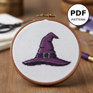 Peut inclure: Un motif de point de croix d'un chapeau de sorcière violet avec une boucle argentée, monté dans un cercle à broder en bois. Le texte "PDF PATTERN" est dans un cercle blanc en haut à droite. Le fond est blanc.