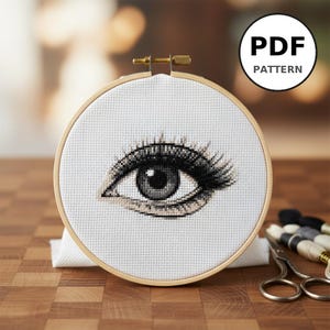 Peut inclure: Une broderie au point de croix d'un œil, dans un cercle en bois. L'œil est rendu en nuances de gris et de noir, avec des cils détaillés. Le cercle est posé sur un fond flou. Le texte "PDF PATTERN" est dans un cercle blanc.
