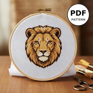 Peut inclure: Un motif de point de croix d'une tête de lion, aux tons bruns et beiges, est présenté dans un cercle à broder en bois. Le texte "PDF PATTERN" est dans un cercle blanc. L'image comprend du fil à broder et des ciseaux.