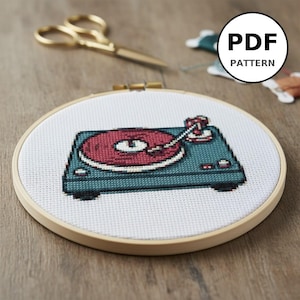 Könnte beinhalten: Ein Kreuzstichmuster eines türkis-roten Plattenspielers wird in einem hölzernen Stickrahmen gezeigt. Der Text "PDF PATTERN" befindet sich in einem weißen Kreis. Goldene Schere und Faden sind im Hintergrund.