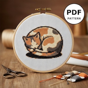Peut inclure: Un motif de point de croix d'un chat calico endormi dans un cercle à broder en bois. Le chat est représenté en orange, marron et crème. Le cercle est posé sur une surface en bois, avec des ciseaux et des bobines de fil. Le texte "PDF PATTERN" est dans un cercle blanc.