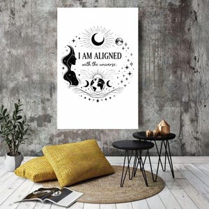 May include: A white canvas print with a black illustration and text. The artwork features a woman's silhouette, crescent moons, stars, and the words "I AM ALIGNED with the universe." The print is displayed in a room with yellow pillows and black side tables.