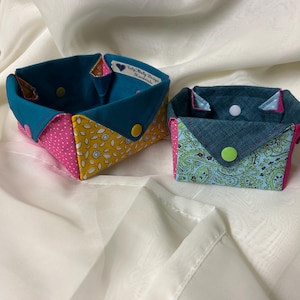 May include: Two handmade fabric baskets. One is teal, yellow floral, and pink polka dot. The other is teal paisley, pink, and teal. Both have button closures. The label on the larger basket reads "Toly-Moly Design Handmade."