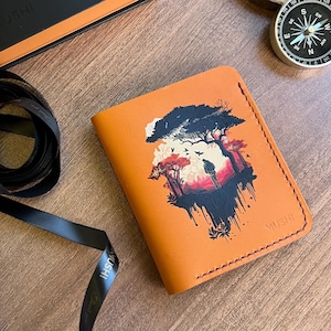 May include: An orange leather wallet with a landscape design, a black gift box, a compass, and a decorative wooden piece are arranged on a wooden surface. A black ribbon and a roll of striped wrapping paper are also present.