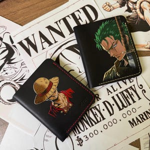 May include: Two black leather wallets featuring anime character illustrations. One wallet displays a character with a straw hat and red shirt, while the other shows a green-haired character with a sword. Both wallets have visible stitching.