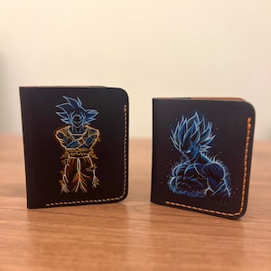 May include: Two black leather wallets with orange stitching. One wallet features a neon blue and orange illustration of a character with spiky hair and crossed arms. The other wallet has a blue illustration of a muscular character.