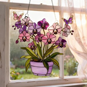 May include: Stained glass orchid window hanging with pink, purple, and white flowers in a lavender pot. Two butterfly accents are attached to the top with chains. The window art is displayed in front of a window with a sheer curtain.