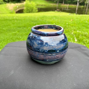 May include: A round ceramic planter with a glossy finish featuring a deep blue and purple mottled glaze. The lower section displays a subtle embossed diamond pattern, while the interior reveals a smooth, light beige surface for planting.