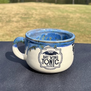 May include: A ceramic mug with a white base and a blue rim and handle. The mug features the text "BAT WING TONIC ELIXIR" in a black and white design. The mug is sitting on a dark surface.