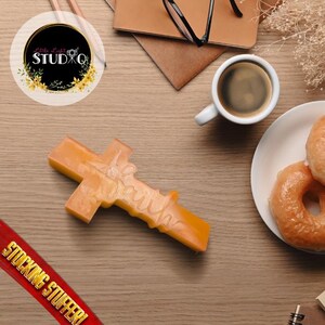 Puede incluir: Una cruz naranja con la palabra "Faith" inscrita. Descansa sobre una superficie de madera, cerca de rosquillas glaseadas y una taza de café. También es visible un letrero rojo que dice "Stocking Stuffer". La cruz es probablemente de cera o jabón.