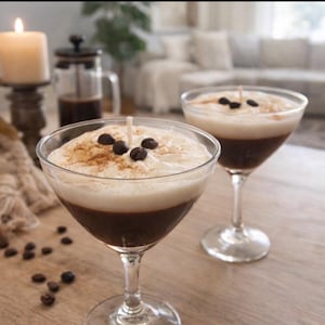 May include: Two cocktail-style candles with a coffee theme. The candles are in clear glasses, with a dark brown base, a white foam top, and topped with coffee beans and cinnamon. A lit candle and coffee press are in the background.