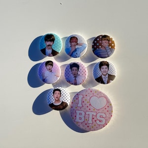 May include: A collection of eight round pinback buttons. Seven buttons feature images of people, and one pink button has the text "I BTS" with a heart. The buttons are on a white surface.