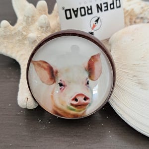 May include: A round pendant with a watercolor painting of a pig's face, set in a brown metal frame. The pig has pink ears and a pink snout. The pendant is next to a white seashell and a starfish.
