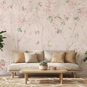 Op de afbeelding: Een woonkamer met een beige bank, kussens en een houten salontafel. Het behang heeft een bloemenpatroon met roze bloesems, vogels en vlinders. Potplanten voegen een vleugje groen toe, wat een rustige sfeer creëert.