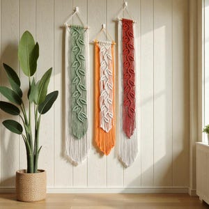 May include: Three macrame wall hangings in green, orange, and red, each featuring a leaf design. The hangings are suspended from wooden dowels and have a fringed bottom. They are mounted on a light-colored wooden wall.
