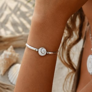 Puede incluir: Una pulsera rígida de tono plateado con un dije circular central que presenta una incrustación de concha en espiral blanca y marrón. La banda metálica es lisa y pulida, conectándose al engaste circular con pequeños detalles esféricos.