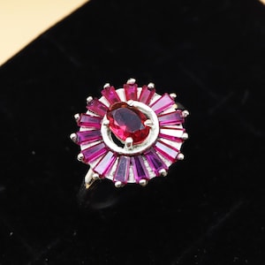 May include: A silver ring featuring a central oval-cut, deep red gemstone surrounded by a sunburst design of smaller, rectangular, magenta-colored stones. The ring is set against a black background.